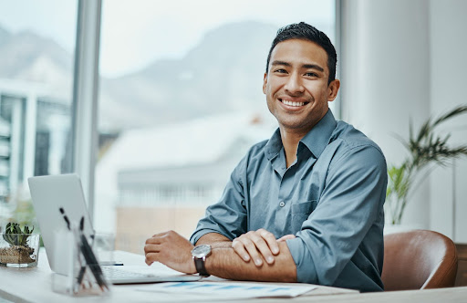 A professional man in a button-down shirt smiles at the camera from his desk